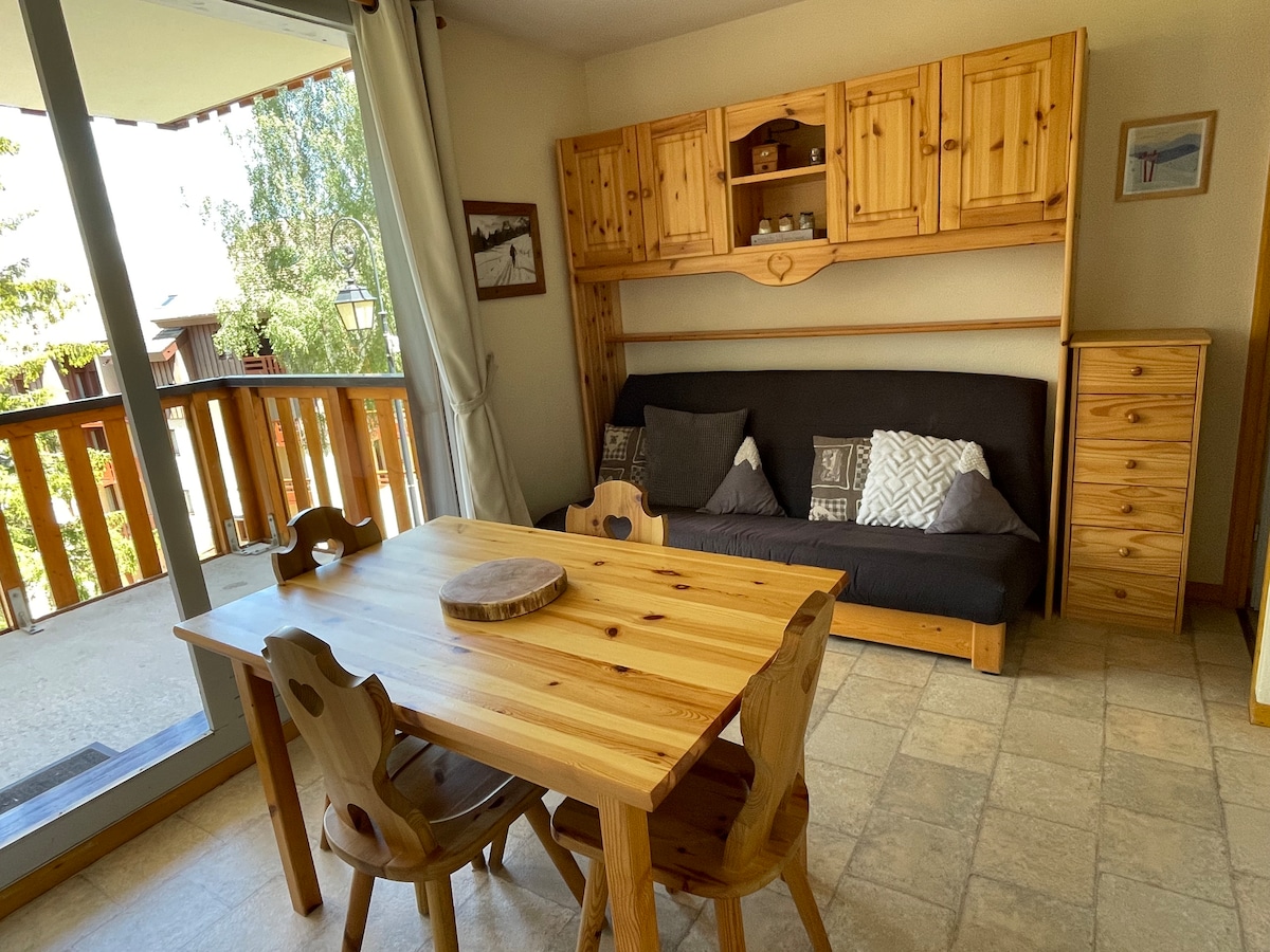 A warm and inviting living area is showcased, featuring a wooden dining table with rounded chairs and a comfortable convertible sofa. Natural light streams through large glass doors that lead to a balcony, enhancing the spacious feel of the room.