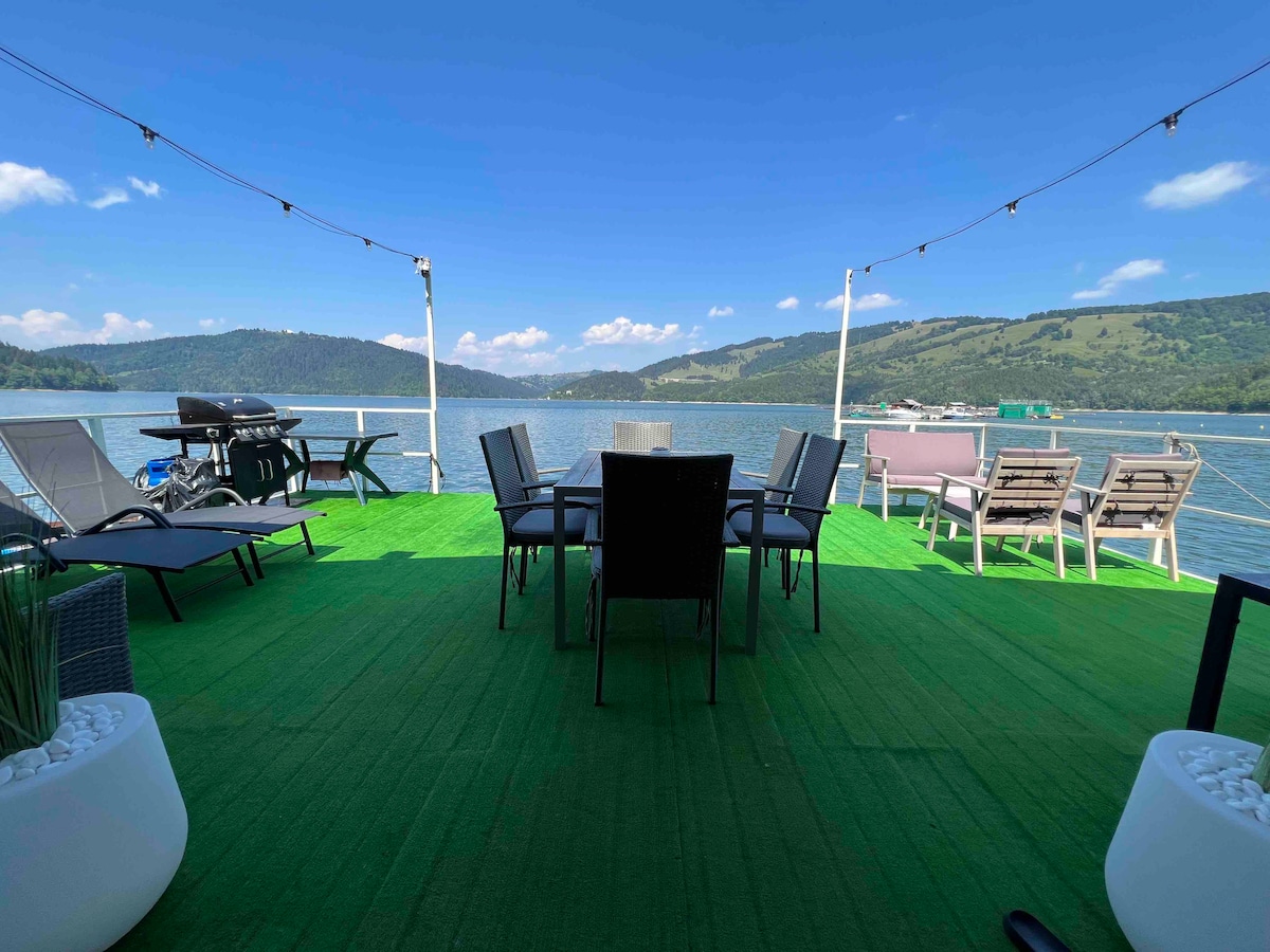 A deck area is presented with green flooring, featuring a dining table surrounded by multiple chairs. Lounge chairs are positioned nearby, with a barbecue grill set off to one side. Scenic views of the lake and surrounding hills are visible under a clear blue sky.