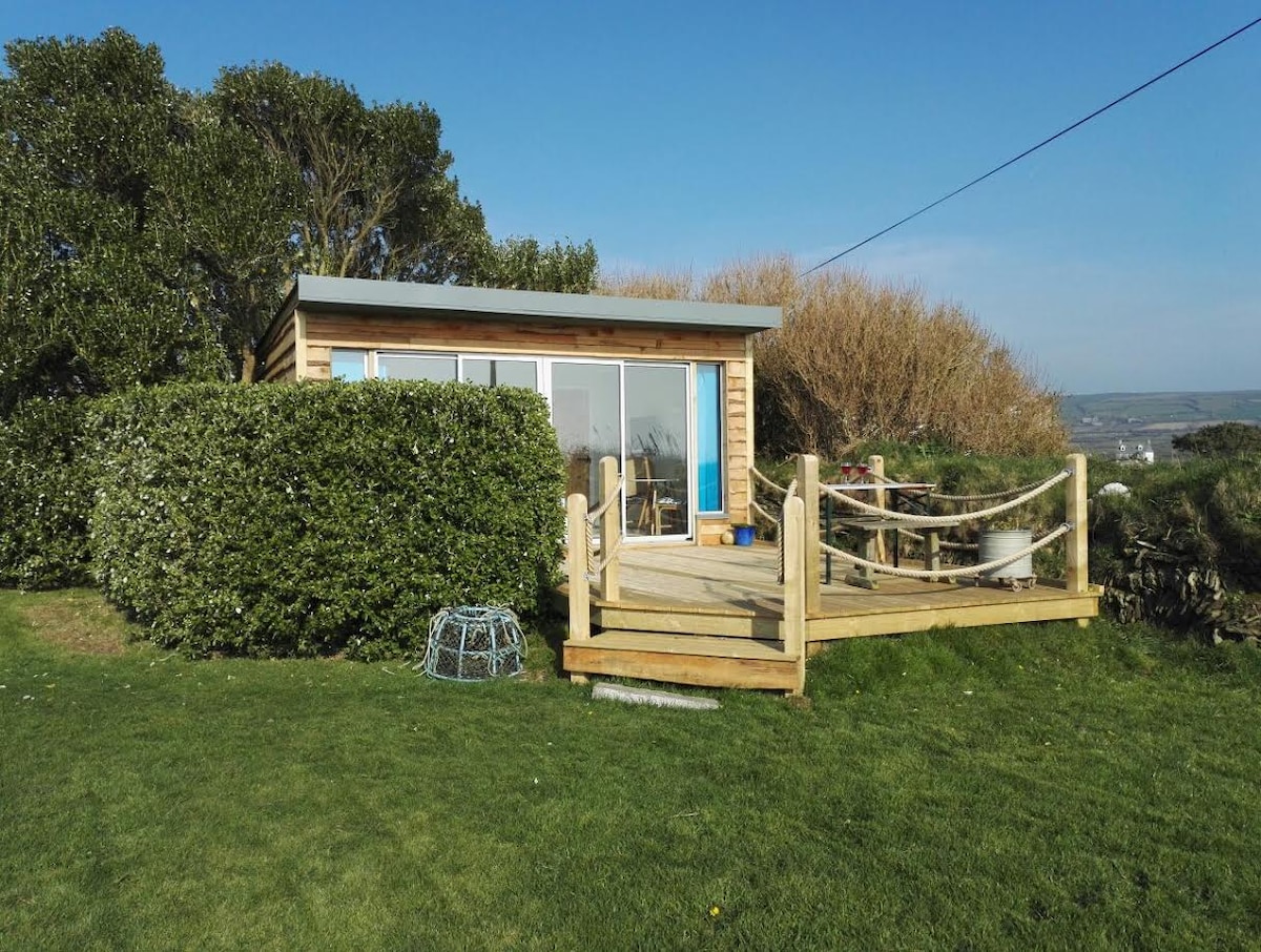 The cabin is nestled within a lush green garden, featuring a wooden deck that extends towards the entrance. Large glass doors open to the outside, allowing natural light to illuminate the interior. A variety of shrubs and trees create a natural boundary, enhancing privacy.