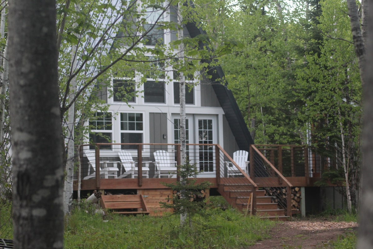 The A-frame cabin is surrounded by lush green foliage, featuring a spacious front deck with several white Adirondack chairs. Large south-facing windows provide a view into the interior, while a wooden staircase leads from the deck down to the natural landscape.