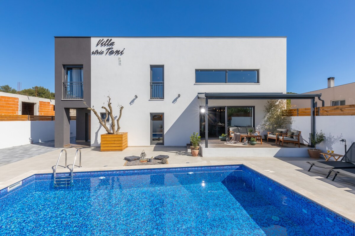 The modern exterior of the villa is highlighted by a striking pool, surrounded by sun loungers and a shaded terrace. The building features large windows, providing a glimpse of the comfortable seating area inside, while a landscaped yard offers additional outdoor space.