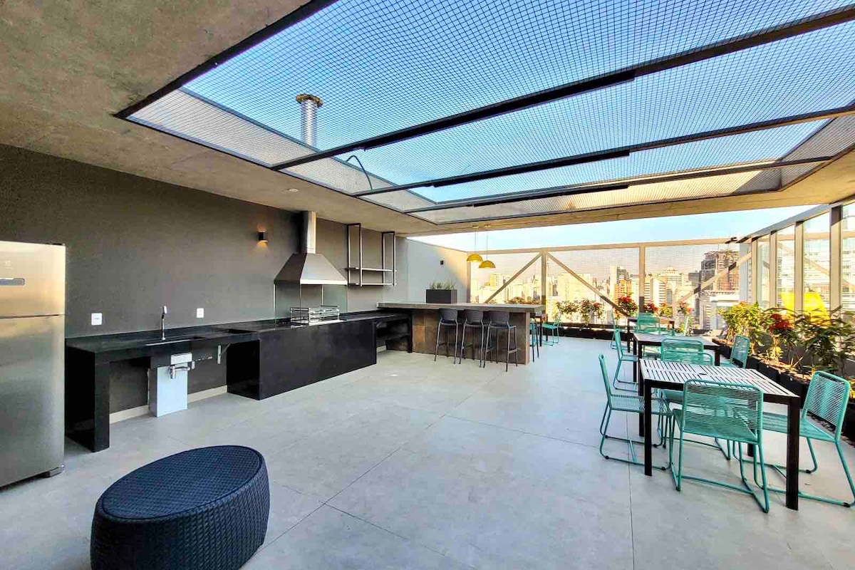 A spacious rooftop area features a modern kitchen with stainless steel appliances, complemented by a large countertop. Multiple seating options include tables and chairs surrounded by greenery. Natural light floods the space through skylights, offering panoramic views of the surrounding city.