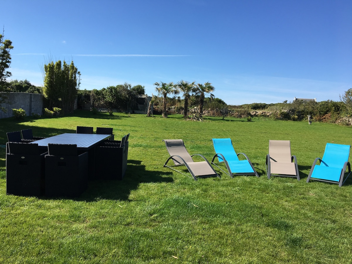 A spacious garden area features a large dining table surrounded by multiple chairs. Several light and dark lounge chairs are arranged on the green lawn under clear blue skies, providing a serene setting for relaxation.