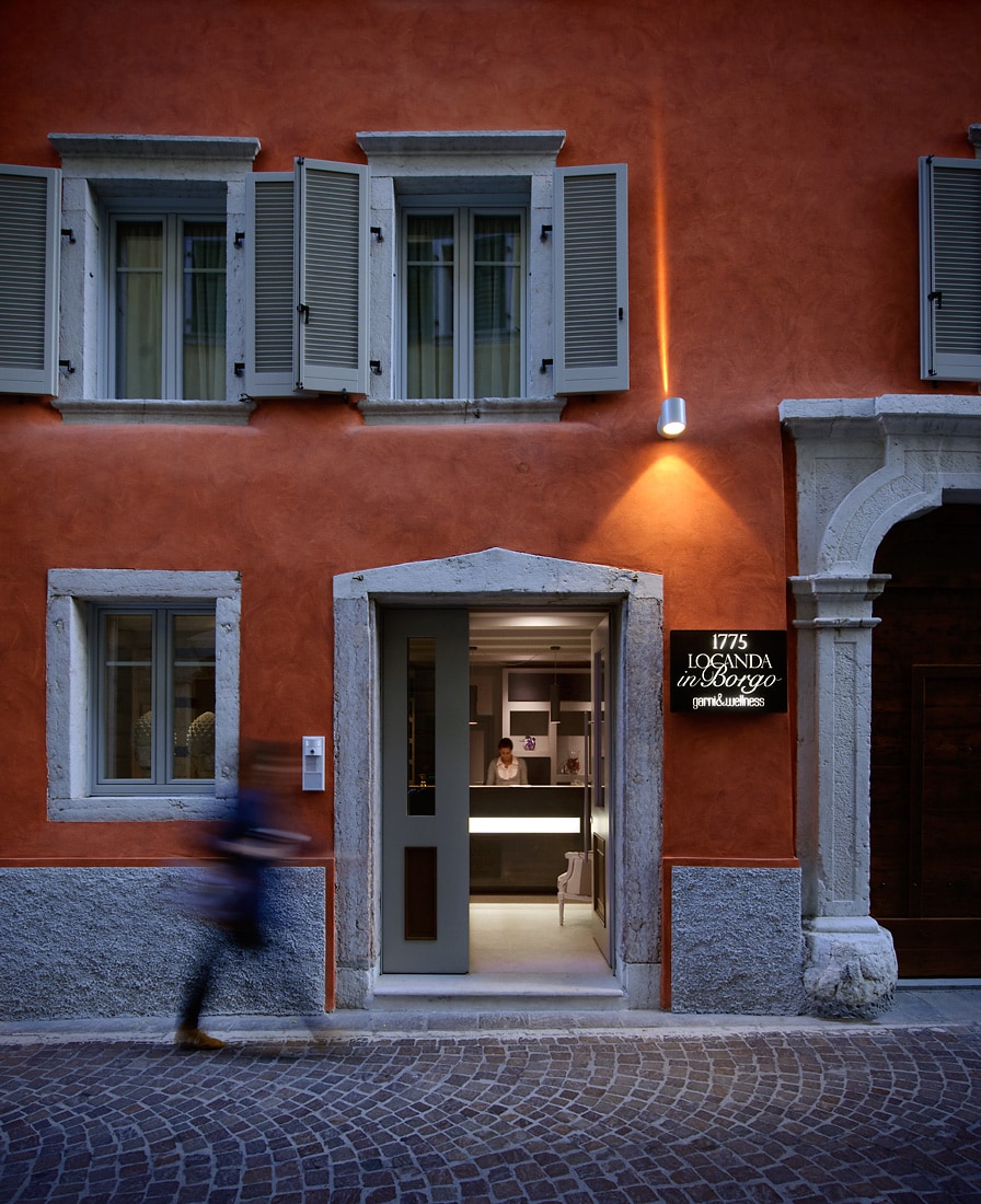 An elegant building with an orange exterior features several light green shutters. The entrance showcases a modern door and sign, while soft lighting highlights the welcoming atmosphere. A cobblestone street runs in front, adding to the quaint charm of the location.