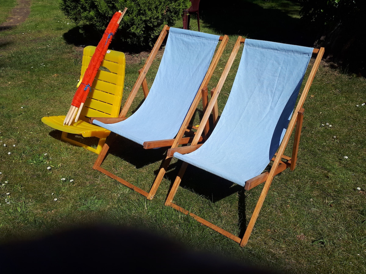 Two deck chairs are positioned on a grassy area, featuring one in a vibrant yellow color and the other in a light blue shade. A colorful sun umbrella rests nearby, adding to the outdoor seating arrangement.