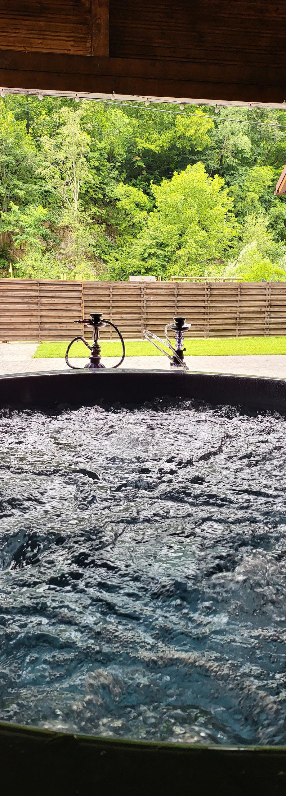 The image captures a hot tub filled with bubbling water, set against a backdrop of greenery. The surrounding area features a wooden fence, providing privacy and a sense of seclusion. Natural light filters through, enhancing the serene environment.