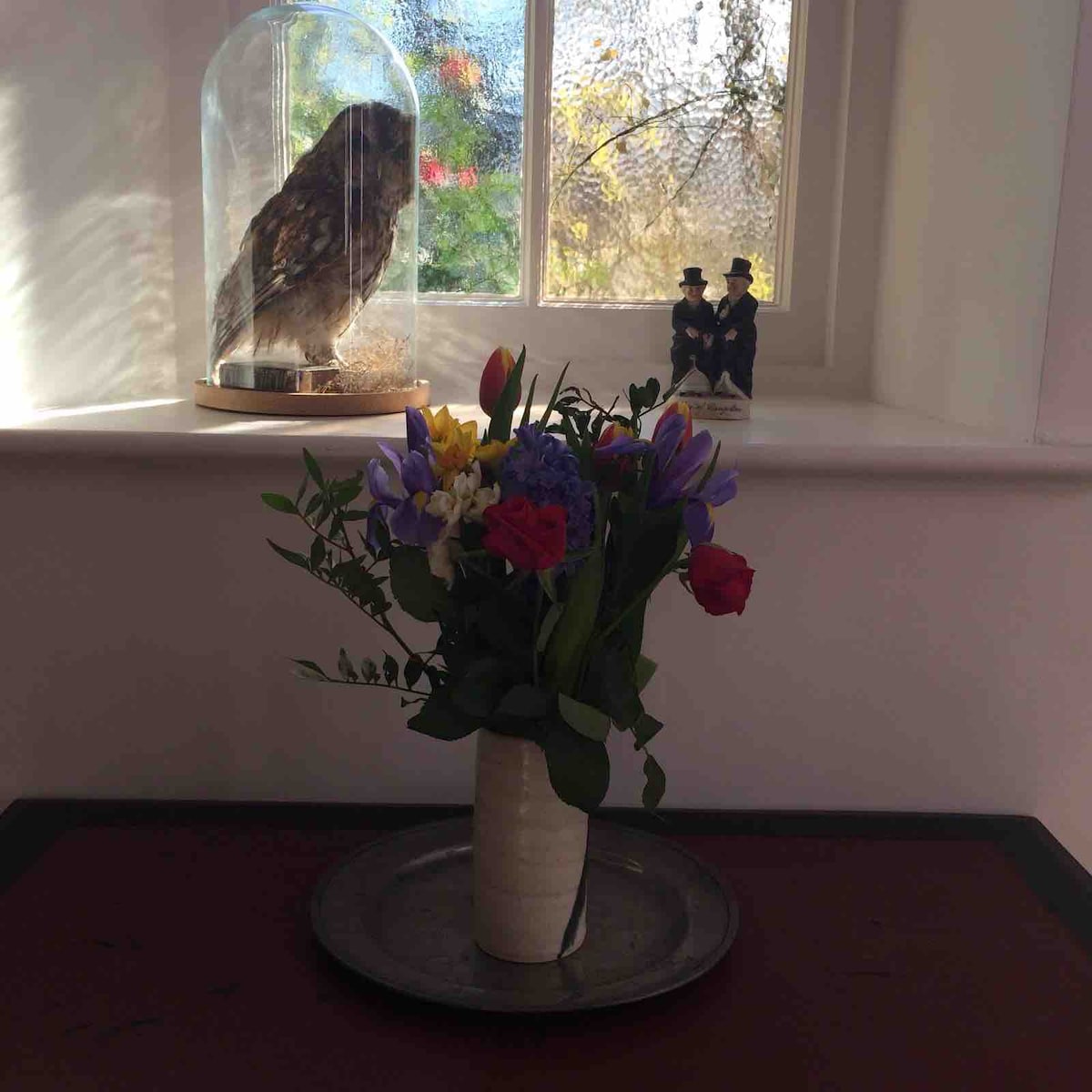 A decorative arrangement features a bouquet of colorful flowers in a simple vase, placed on a metal tray. Behind it, a glass display contains a mounted owl, accompanied by a pair of miniature figurines. Natural light filters through the nearby window.