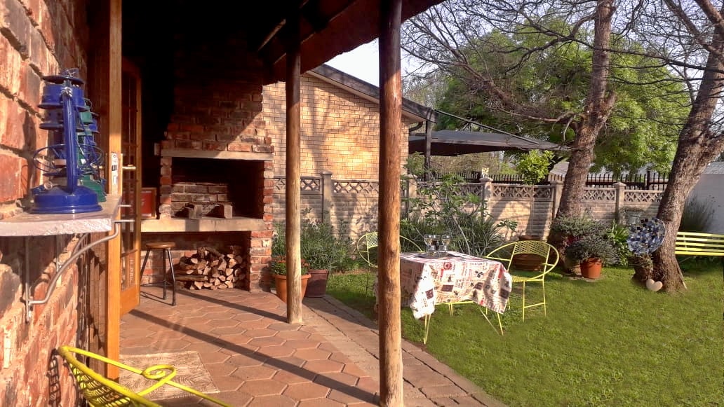 A covered patio features a built-in braai area with stacked firewood nearby. Garden furniture, including a small round table with a floral tablecloth and two chairs, is positioned on the green lawn, surrounded by potted plants and trees.