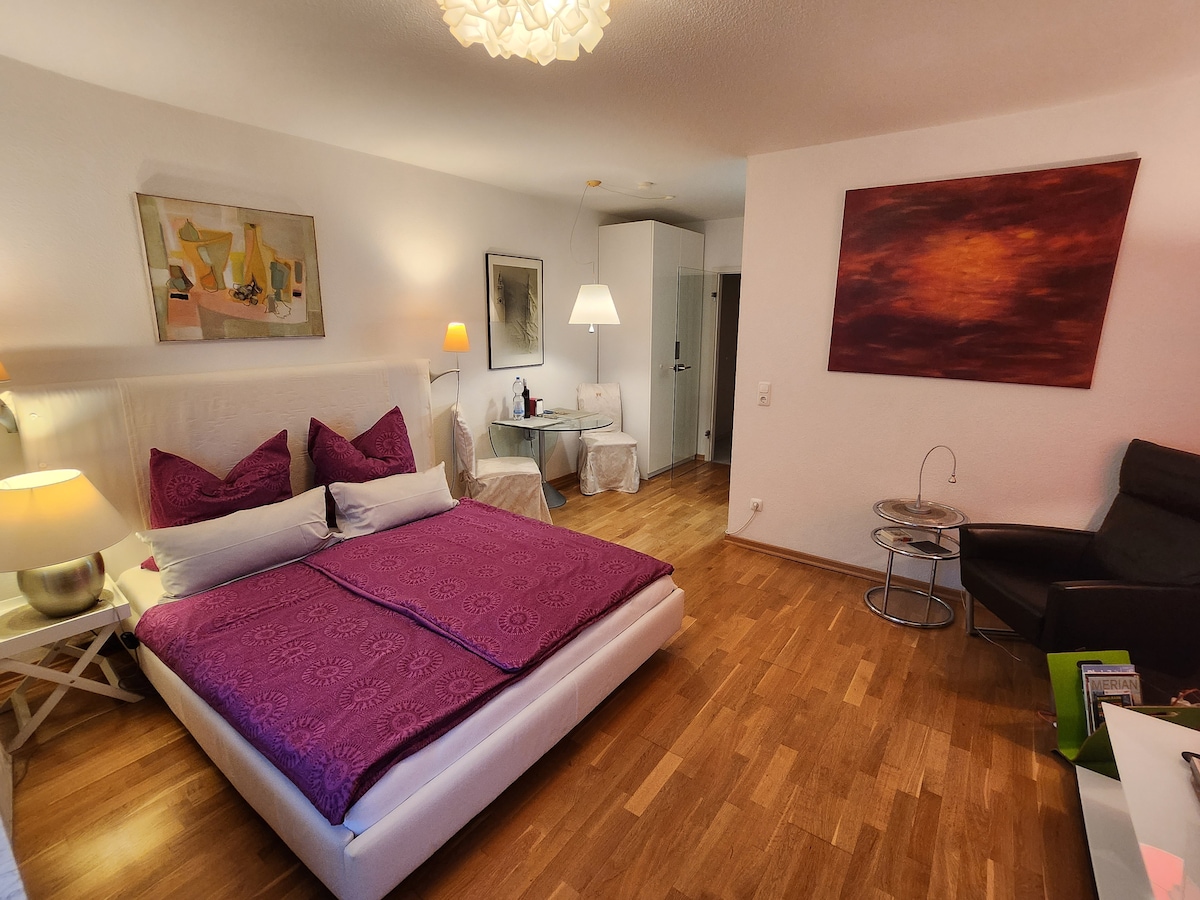 A spacious bedroom showcases a large white bed adorned with a deep purple bedspread and coordinating throw pillows. Complementary lamps provide gentle lighting, while minimalist furniture includes a stylish armchair and a side table. A colorful wall art piece adds visual interest to the neutral decor.