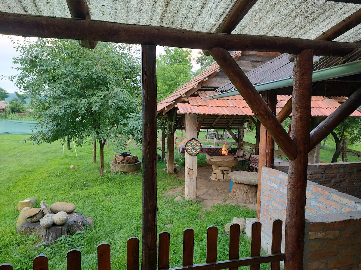 A spacious outdoor area showcases a covered seating space with log furniture and a stone table. A fire pit is raised on stone, surrounded by grassy areas and trees. A rustic wooden fence frames the view, enhancing the natural surroundings.