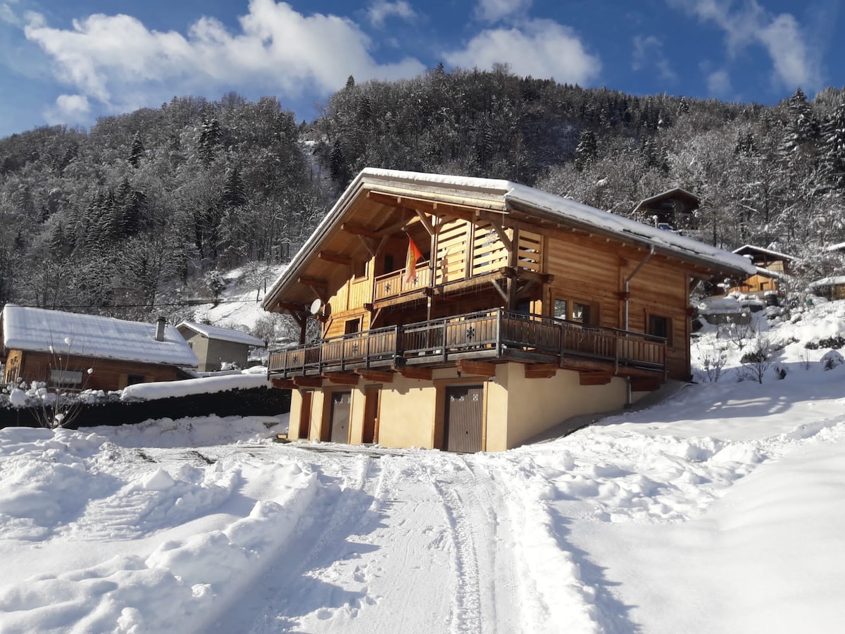 A wooden chalet is nestled in a snowy landscape, showcasing its sloping roof and front balcony. The exterior features warm wood tones against a backdrop of trees and hills covered in snow. A snow-covered driveway leads to the garage below.