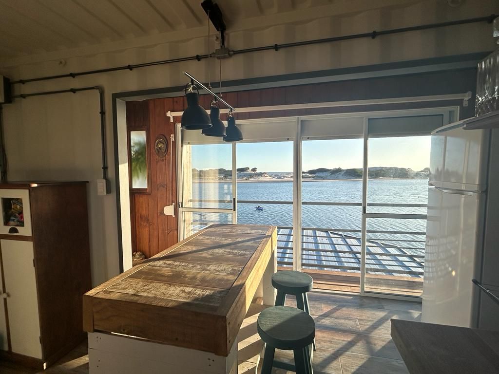 An open kitchen area features a large wooden table surrounded by green stools. Floor-to-ceiling sliding glass doors reveal a serene view of the water, inviting natural light into the space. The scene includes a glimpse of the outdoor deck, enhancing the connection with nature.