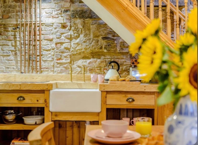 A rustic kitchen area features natural wood cabinetry, including a spacious farmhouse sink. Exposed stone walls add character, while a kettle and dishware suggest a welcoming space for meal preparation. A bouquet of sunflowers is displayed in a vase, adding a touch of warmth to the scene.