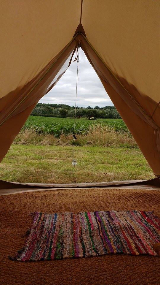 The interior of the tent features a colorful woven rug on the floor, with the entrance offering a view of the grassy meadow beyond. A gentle landscape of fields and greenery is visible through the open tent flap, enhancing the camping atmosphere.