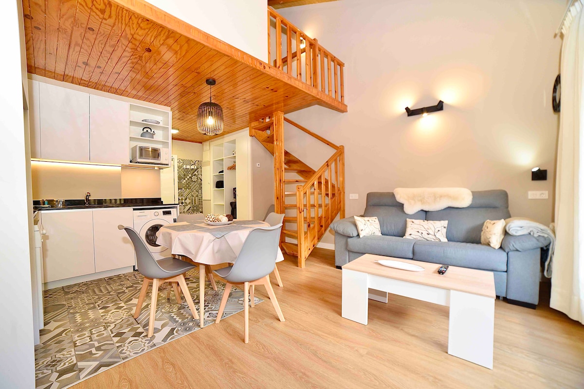 An open-plan living space features a light, airy atmosphere with wooden beams and a light wood floor. A cozy sofa sits across from a white coffee table. A dining table is set with four chairs, and a kitchenette is visible, equipped with modern appliances.