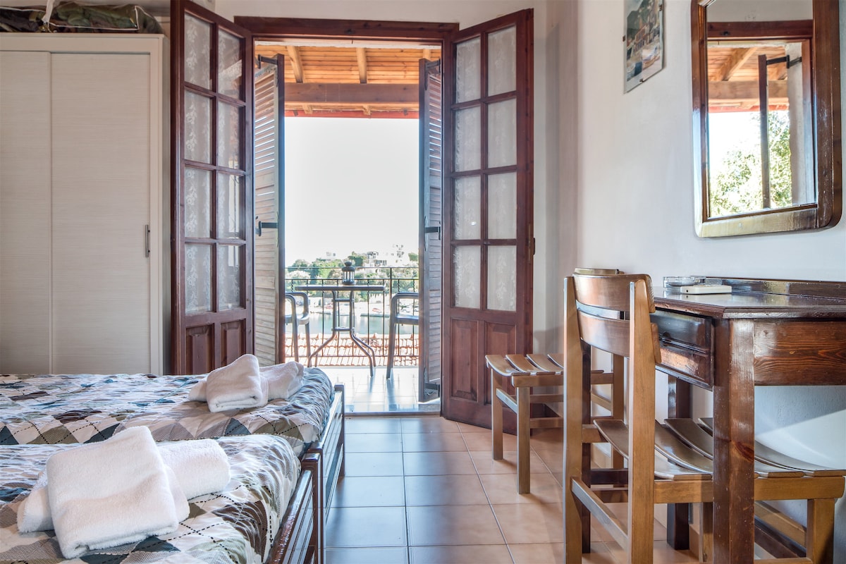 The interior space features twin beds with neutral bedding, a small wooden desk, and a chair. A folding table is positioned near an open balcony door that offers a view of the outdoors. Natural light fills the room, enhancing the warm atmosphere.