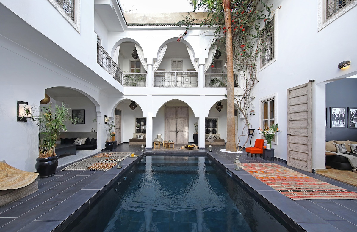 An inviting courtyard is centered around a tranquil pool, surrounded by archways and greenery. Comfortable seating areas with neutral-toned cushions are positioned along the walls, complemented by traditional rugs and decorative lighting. Natural light floods the space, creating a serene atmosphere.
