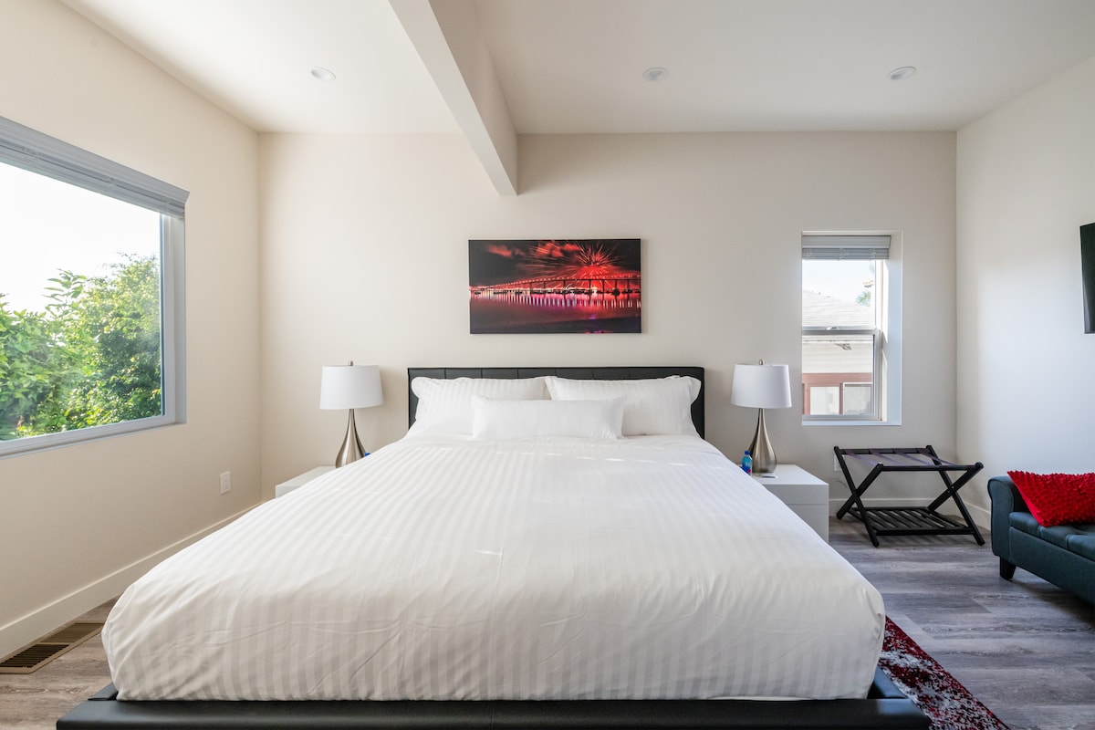 A spacious bedroom is furnished with a large bed covered in a white duvet, flanked by two nightstands with table lamps. A vibrant piece of wall art provides a pop of color above the bed. Large windows allow natural light to fill the room.