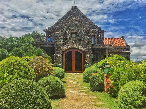 A traditional stone house is framed by a well-maintained garden, featuring rounded hedges and vibrant foliage. The entrance is highlighted by large wooden doors, welcoming guests into the homestay while a bright blue sky and clouds provide a picturesque backdrop.