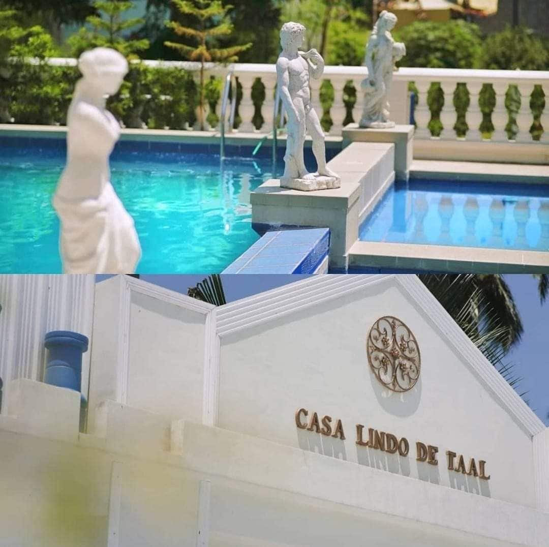 A serene infinity pool reflects the surrounding greenery, featuring elegant statues placed along the edge. The venue name, 'Casa Lindo de Taal,' is prominently displayed on a well-maintained white facade, ensuring visibility and charm. The inviting space is enhanced by a bright and sunny ambiance.