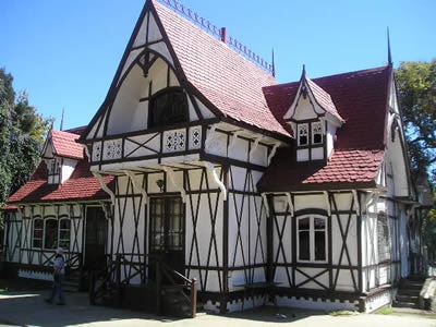 The house features a distinctive architectural style with a combination of white walls and dark wooden beams. A red-tiled roof adds character to the charming structure, which is surrounded by greenery. Multiple windows allow ample natural light to illuminate the interior.