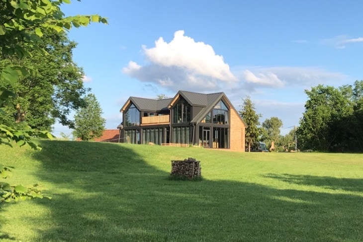 A modern villa is set amidst a lush green landscape, featuring large glass windows that reflect the sky. The structure is complemented by a sloped roof and is surrounded by well-maintained grass, enhancing its tranquil surroundings.
