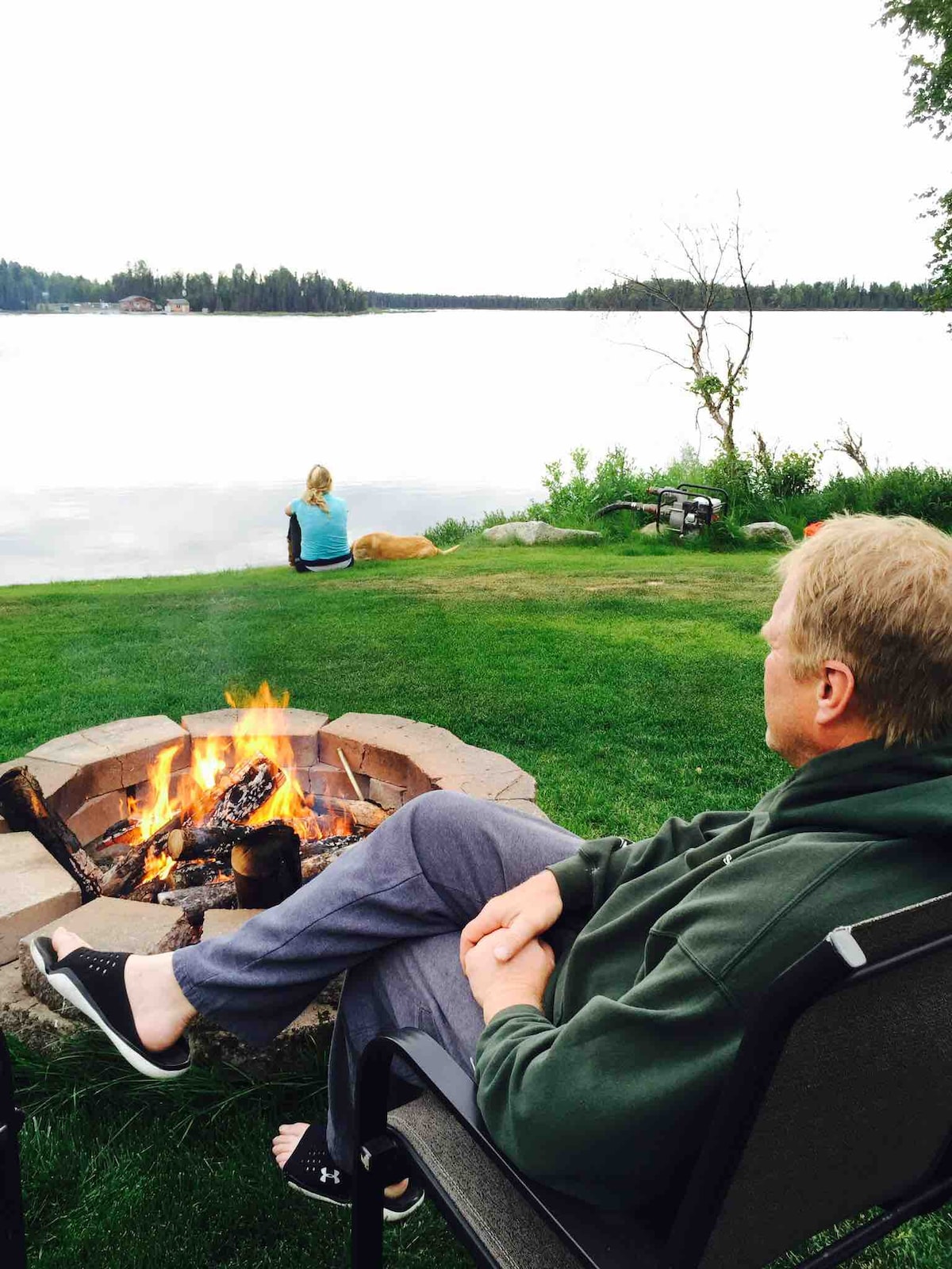 A fire pit made of stone is centrally located, with flames dancing above the logs. In the background, a tranquil lake reflects surrounding greenery, while a person sits quietly on the grass. Another individual enjoys the view from a distance, indicating a peaceful outdoor gathering.
