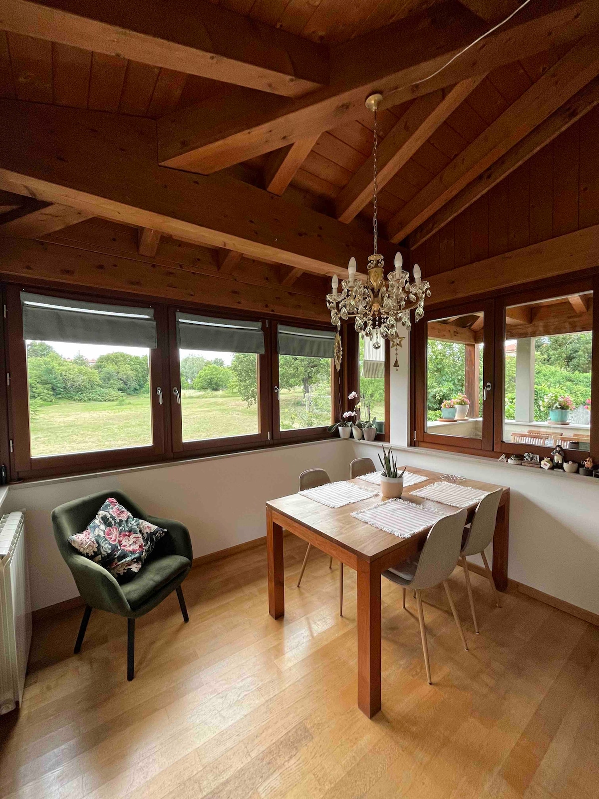 A dining area showcases a wooden table surrounded by four chairs. Large windows frame views of the garden, inviting natural light into the space. A chandelier hangs from the beamed ceiling, adding an elegant touch to the warm interior. Decorative items can be seen on the windowsill.
