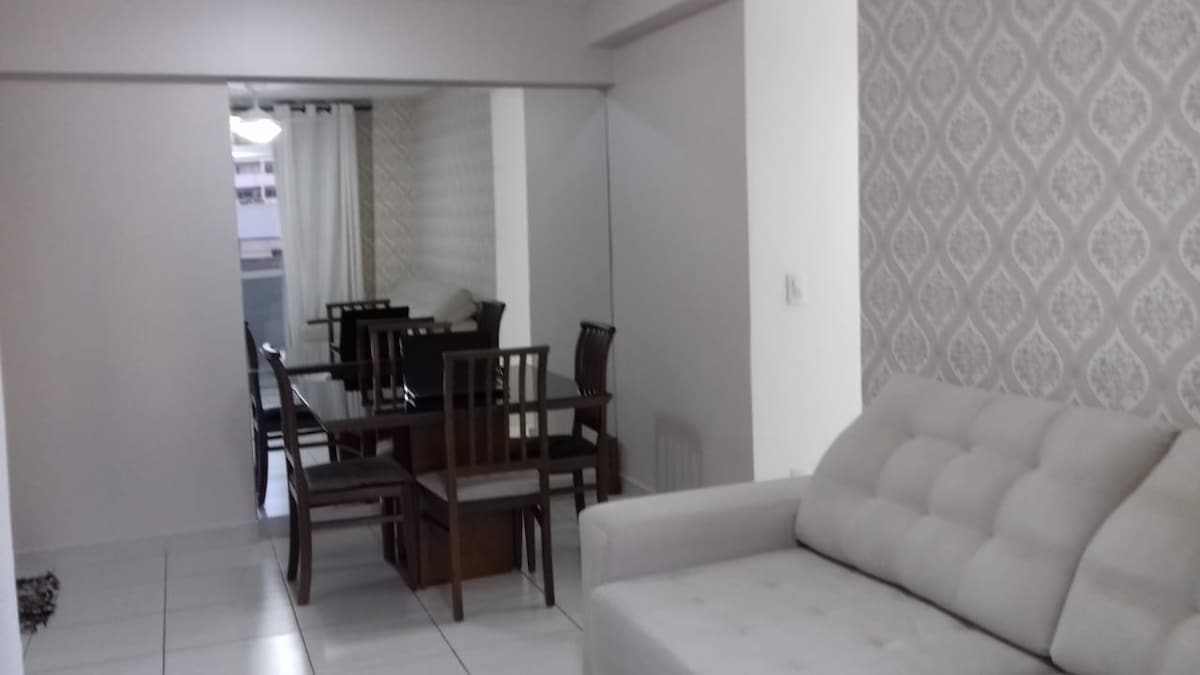 A light-filled living area includes a comfortable sofa and a dining space with a wooden table surrounded by chairs. A large mirror extends along one wall, creating an illusion of spaciousness. Subtle wallpaper adds texture to the neutral-toned room.