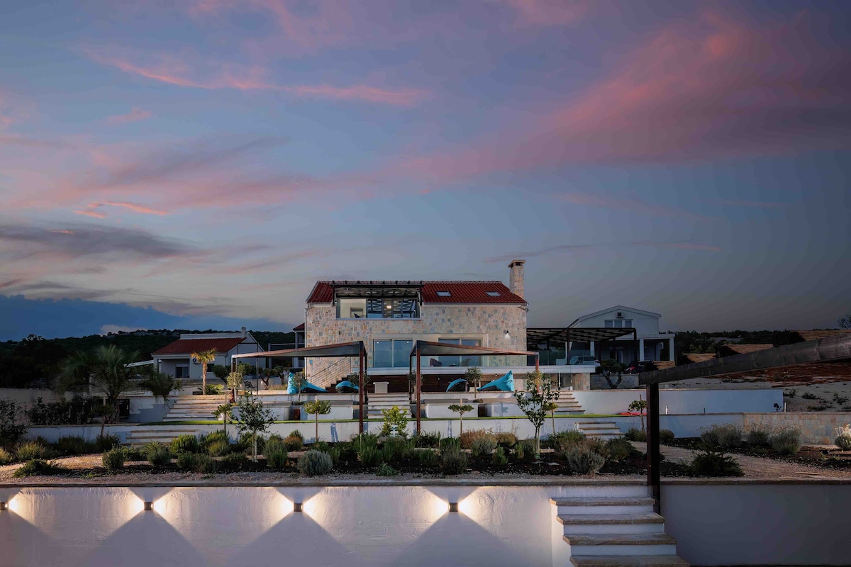 A modern villa with a stone exterior and a red-tiled roof is set against a twilight sky. Lush greenery and neatly arranged outdoor spaces are highlighted in the foreground, with soft outdoor lighting illuminating the pathway leading to the entrance.