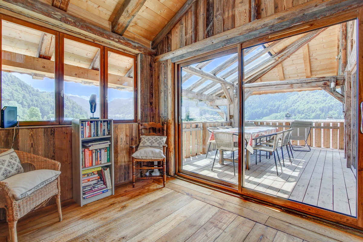 A spacious open plan living area is shown with large glass sliding doors that open to a sunlit terrace. Wooden beams and rustic decor characterize the space, featuring a cozy reading nook with a bookshelf and a wicker chair. Scenic views of the village and mountains are visible through the windows.