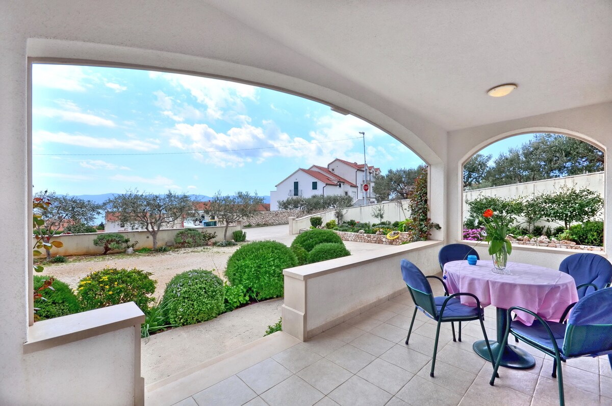 A spacious covered terrace offers seating for four, with a pink table set against a backdrop of a well-maintained garden. Lush green shrubs and flowering plants can be seen, with the sea visible in the distance, framed by an archway that enhances the outdoor view.