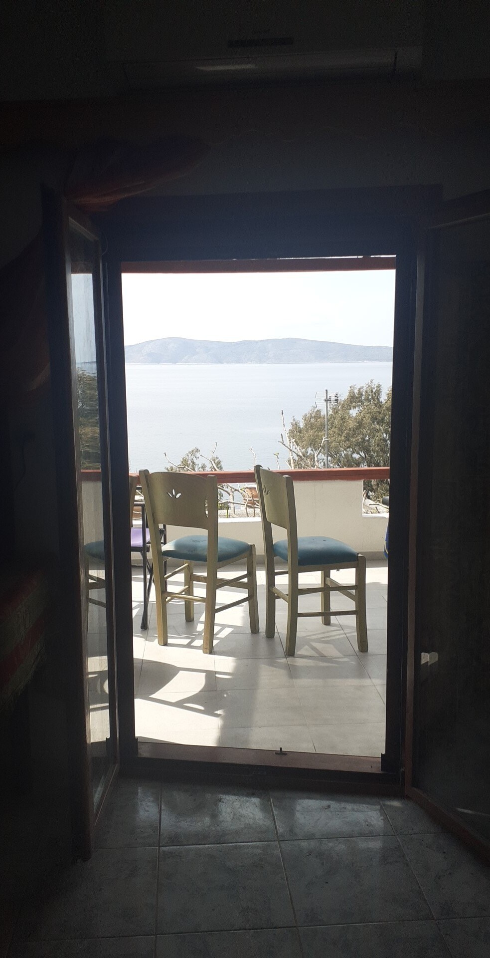 A doorway frames a view of a balcony with three chairs arranged around a small table. The serene sea and distant mountains are visible through the open door, creating an inviting transition between indoor and outdoor spaces.