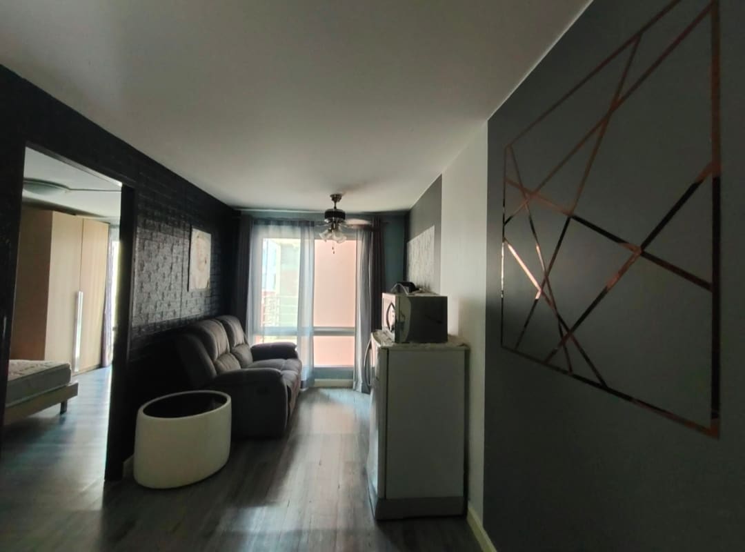 A stylish living area is highlighted by a comfortable dark sofa facing a modern entertainment unit. A minimalist white planter sits on the floor, while a decorative wall art piece with geometric patterns adds interest. Soft natural light enters through large windows covered by sheer curtains.