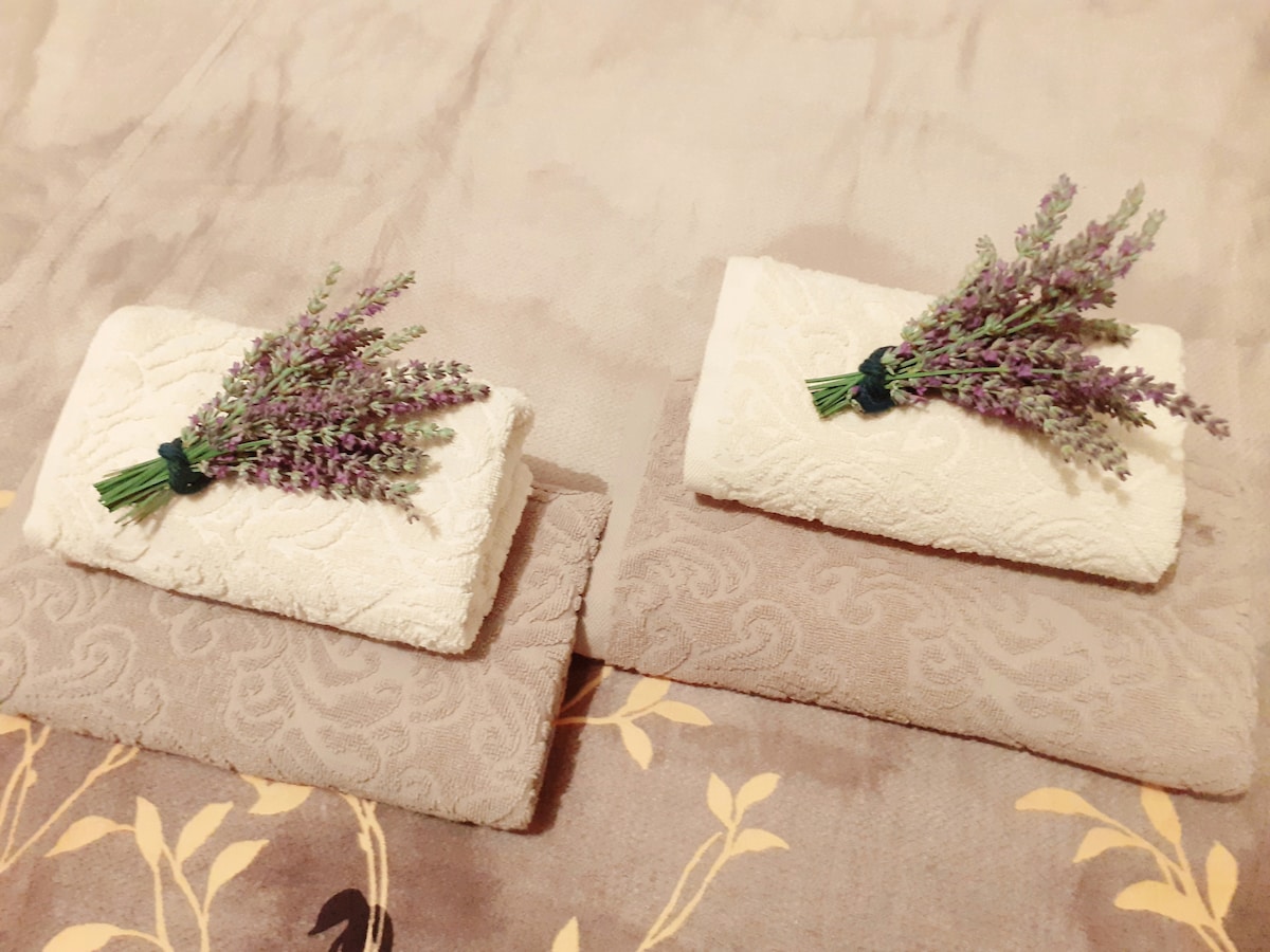 Two rolled white towels are placed neatly on a soft beige surface, each adorned with a sprig of lavender for a touch of decor. The towels are evenly folded, creating a welcoming display.