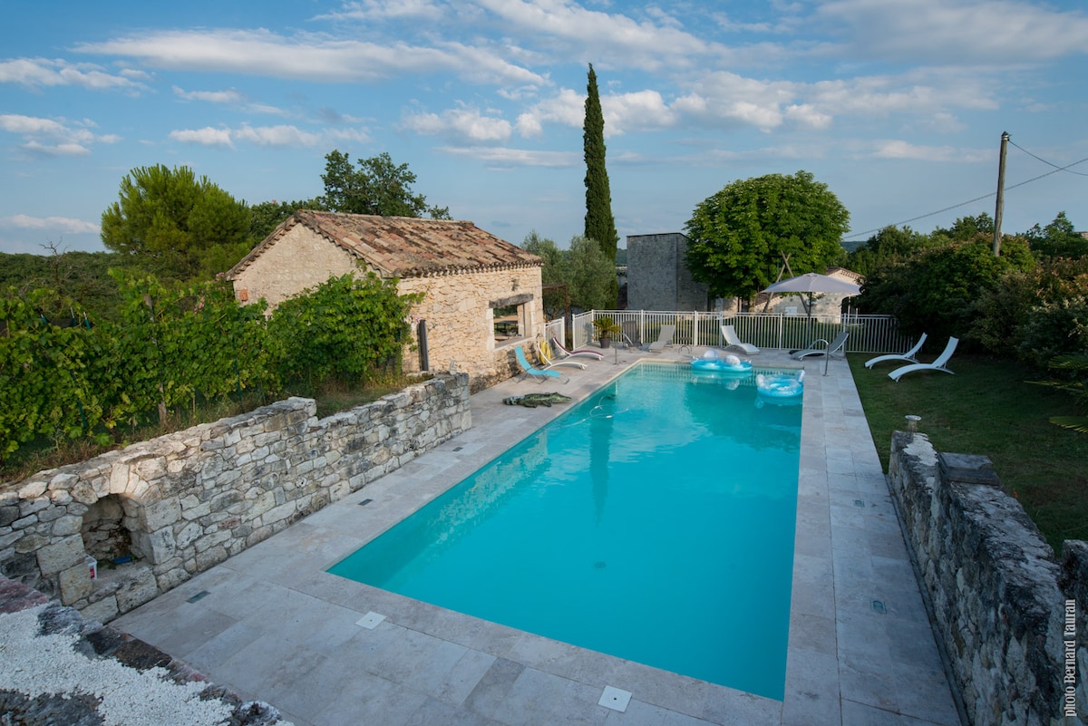 A large, inviting swimming pool is set within a serene landscape, surrounded by lush greenery and rustic stone walls. Sun loungers are placed alongside the pool, offering a relaxing area. The sky is clear with soft clouds, enhancing the tranquil atmosphere.