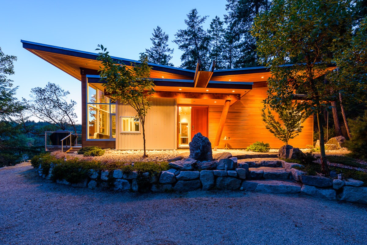 The guesthouse exterior is showcased at twilight, featuring a distinctive butterfly roofline. Warm lighting illuminates the wooden facade, while surrounding greenery and a stone pathway lead to the entrance, enhancing the tranquil forest ambiance.