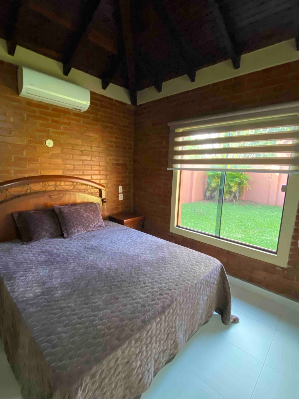 A cozy bedroom features a spacious bed with a quilted cover, positioned against a brick wall. Natural light enters through a large window with blinds, offering a view of the green backyard. An air conditioning unit is mounted above for comfort.