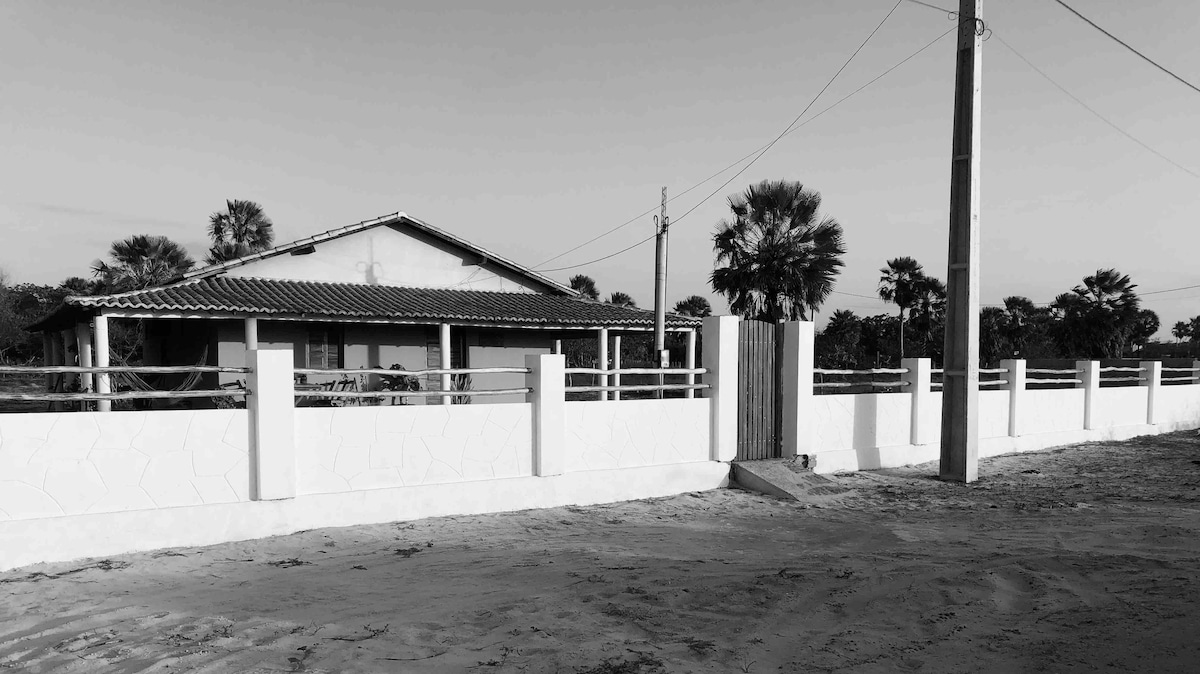 An exterior view of a house is presented, featuring a light-colored facade and a tiled roof. A white fence encircles the property, with a metal gate partially open. Palm trees are visible in the background, providing a hint of greenery amidst the sandy landscape.