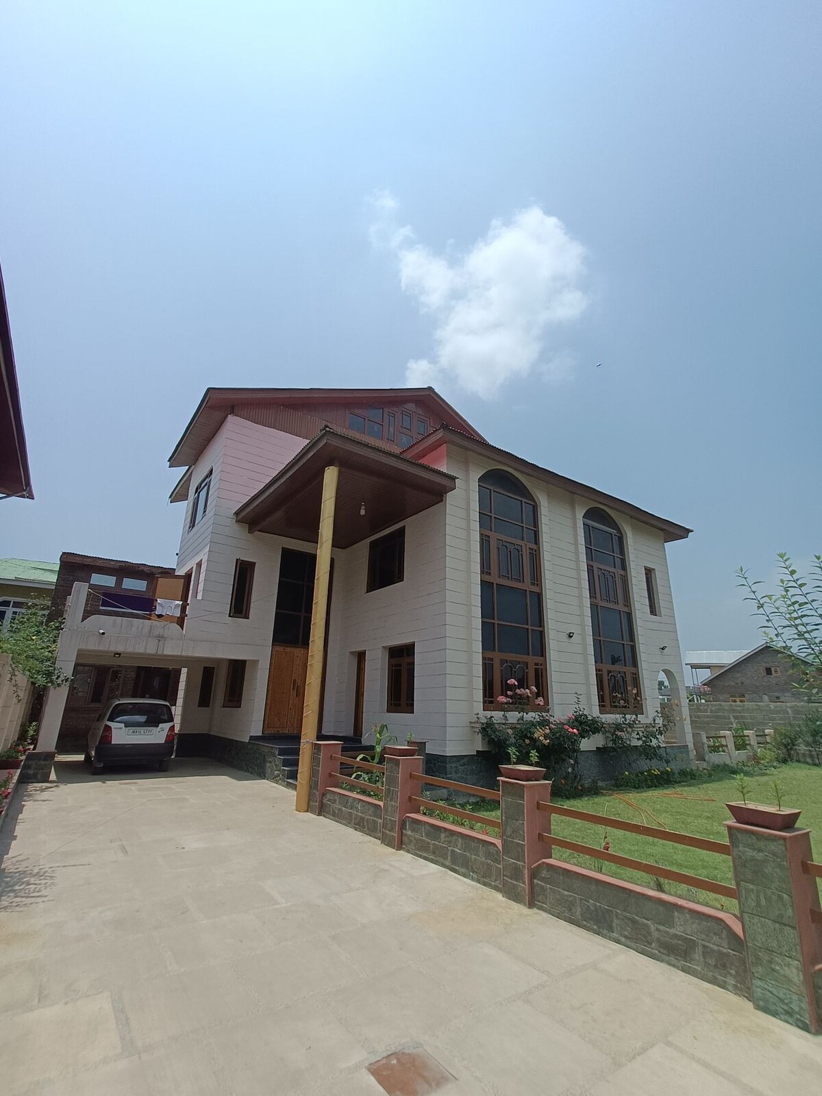 The exterior of the two-story house features large arched windows, allowing ample natural light. A well-maintained garden surrounds the home, with shrubs and paved pathways. A car is parked in the driveway, and the structure is complemented by a clear blue sky.