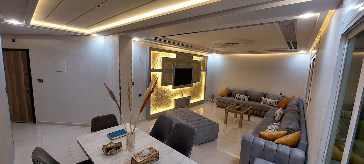 A modern living area showcases a spacious sectional sofa with accent pillows and a lighted feature wall. A dining table is set nearby, complemented by decorative items, while soft lighting enhances the room's inviting ambiance. Large windows allow natural light to fill the space.