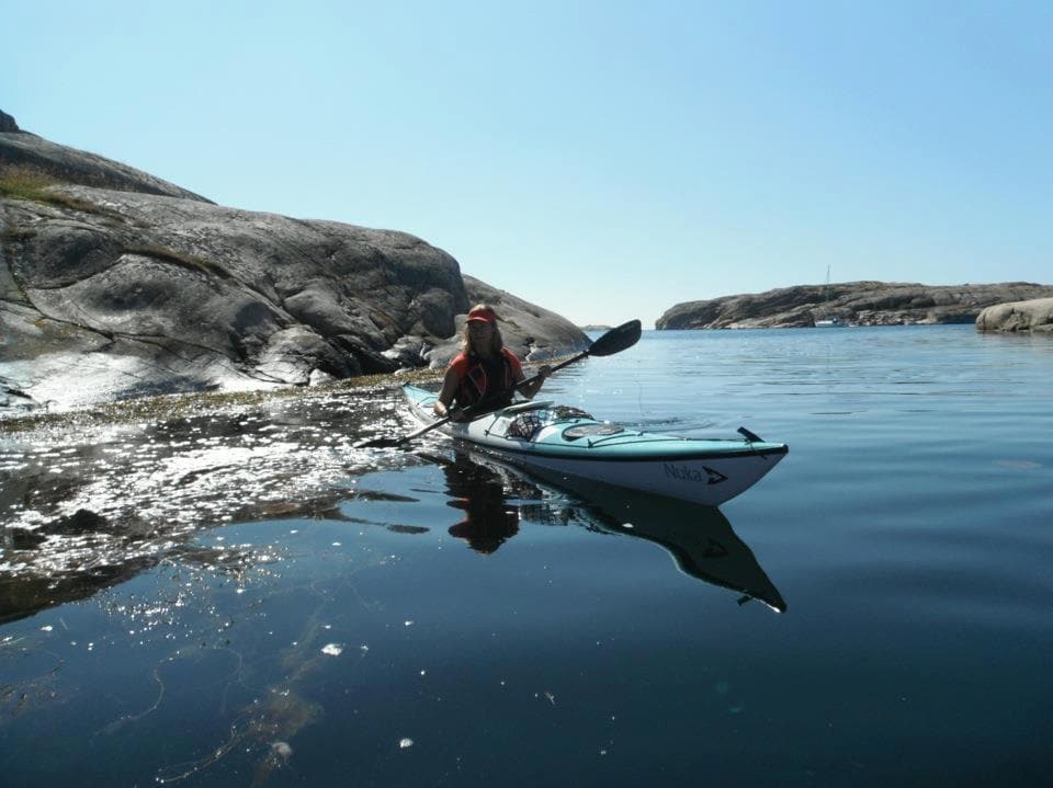 A kayaker is seen paddling in calm waters surrounded by rocky formations and distant islands. The clear blue sky reflects on the water's surface, highlighting the serene coastal environment ideal for kayaking.