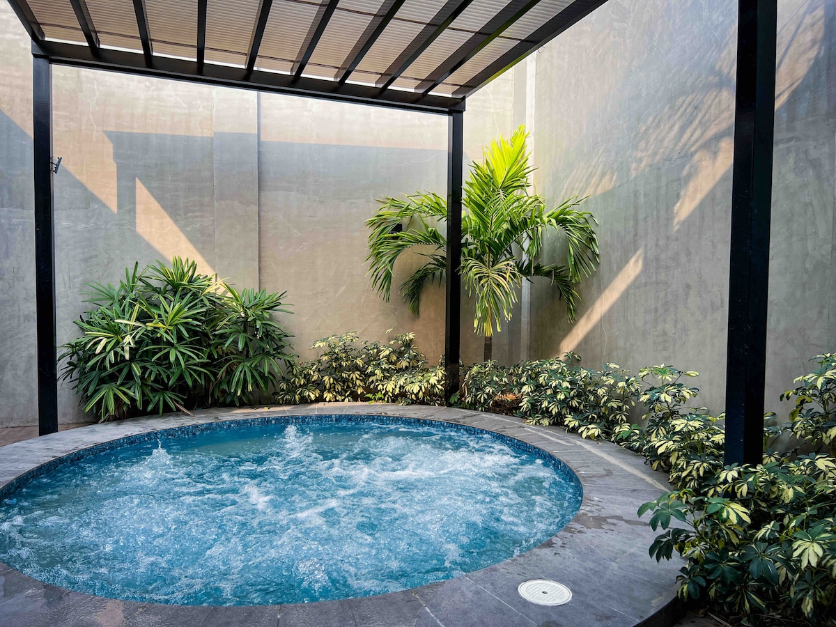 A mini pool is surrounded by tropical plants and decorative foliage. The water is agitated, creating bubbles, while the pool area is shaded by a slatted roof, allowing filtered sunlight to illuminate the space.