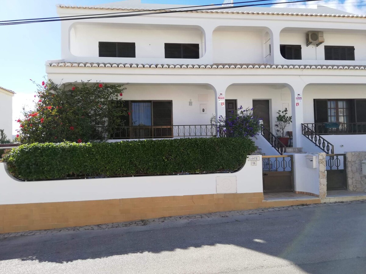 The exterior of the two-story villa features a white façade with dark trim. A balcony with decorative railings is visible on the upper level, framed by vibrant plants. A well-maintained hedge runs alongside the entrance, leading to a set of steps that provide access to the property.