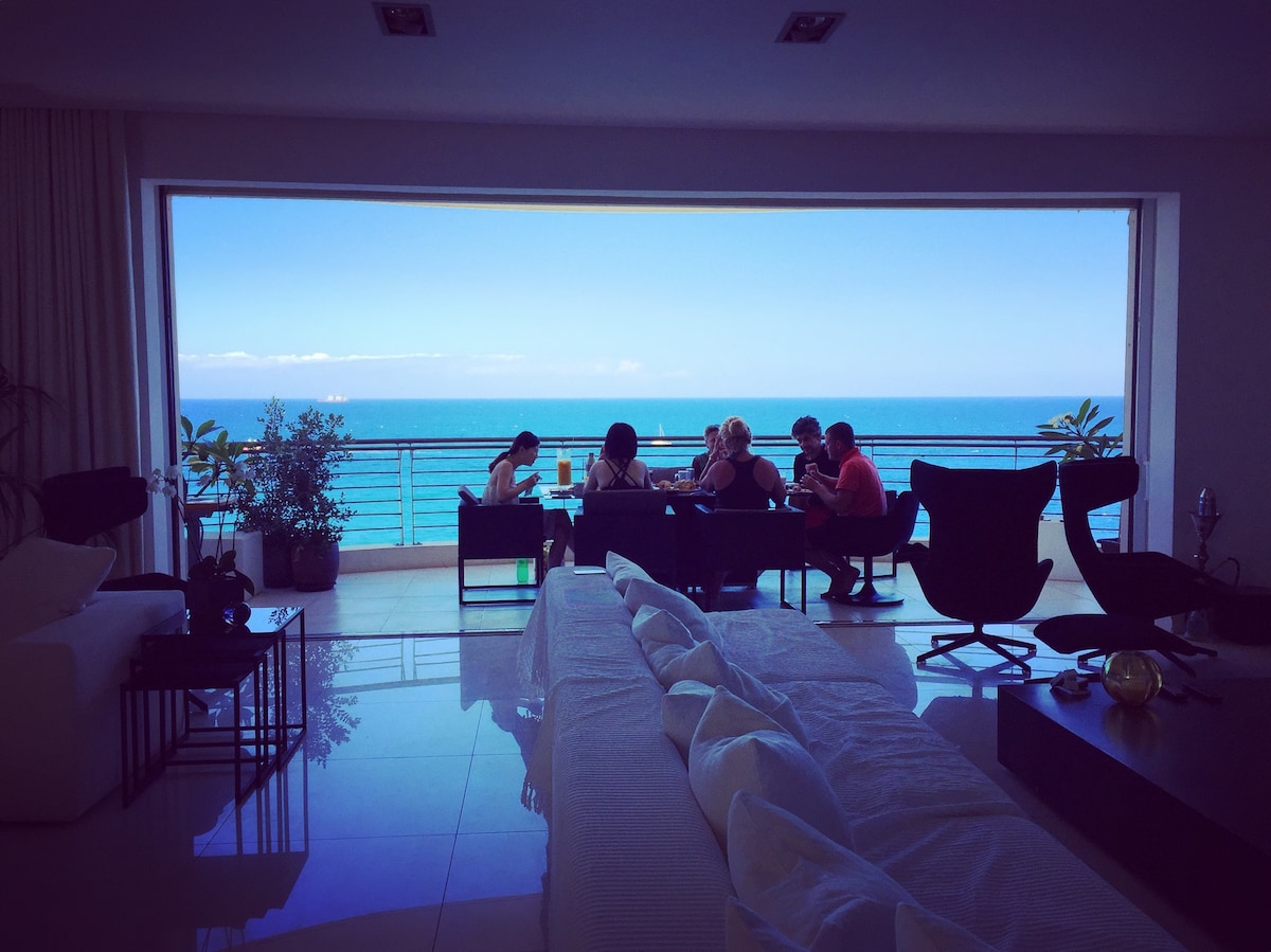 A spacious living area features large glass doors that open to a balcony overlooking unobstructed sea views. Guests are seated at a dining table, enjoying a meal. The room includes comfortable seating arrangements and elements of greenery that soften the modern surroundings.