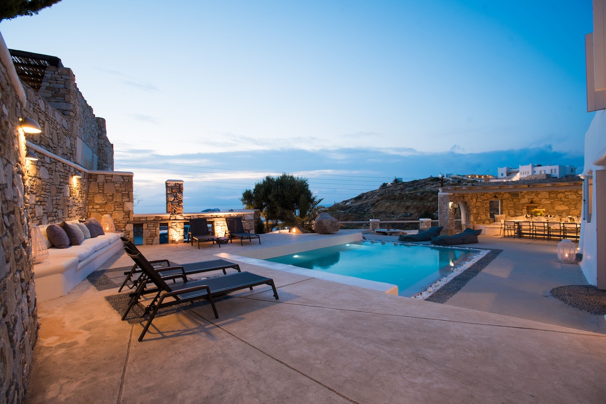 An outdoor area features a private swimming pool surrounded by stone pavers. Sun loungers are arranged near the pool, with comfortable seating visible against a backdrop of hills and the twilight sky. Subtle lighting illuminates the space, enhancing the tranquil evening atmosphere.