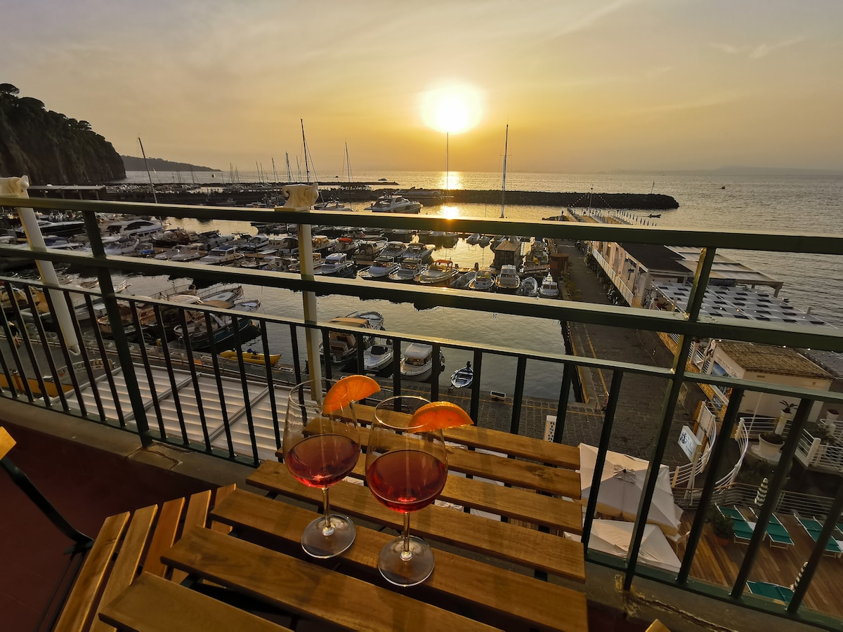 A sunset view is captured from a balcony overlooking the Marina di Cassano, where boats are docked in the harbor. Two glasses of brightly colored drinks are placed on a wooden table, complemented by the warm orange hues of the setting sun reflecting on the water.