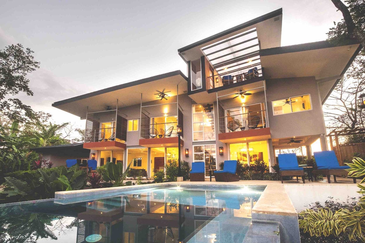 The exterior of the multi-level house features large glass doors and a modern roof design, enhancing the connection with the surrounding nature. A tranquil infinity pool reflects the evening sky, while comfortable lounge chairs line the pool area amidst lush tropical greenery.