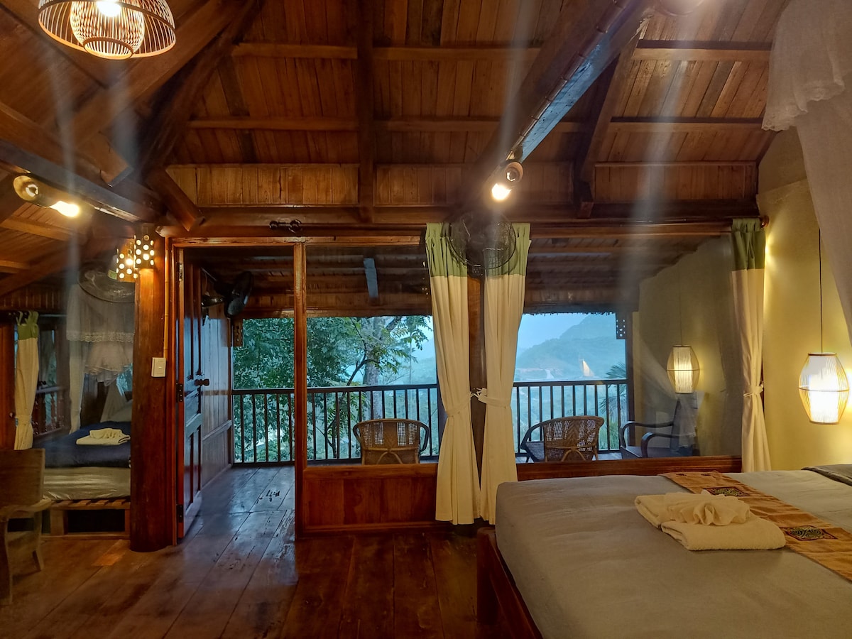 A spacious room features wooden beams and a high ceiling. Natural light filters through large windows, illuminating cozy furniture, including woven chairs. A small balcony overlooks the natural landscape, providing a serene connection to the outdoors. Soft linens adorn the bed, contributing to a calming environment.