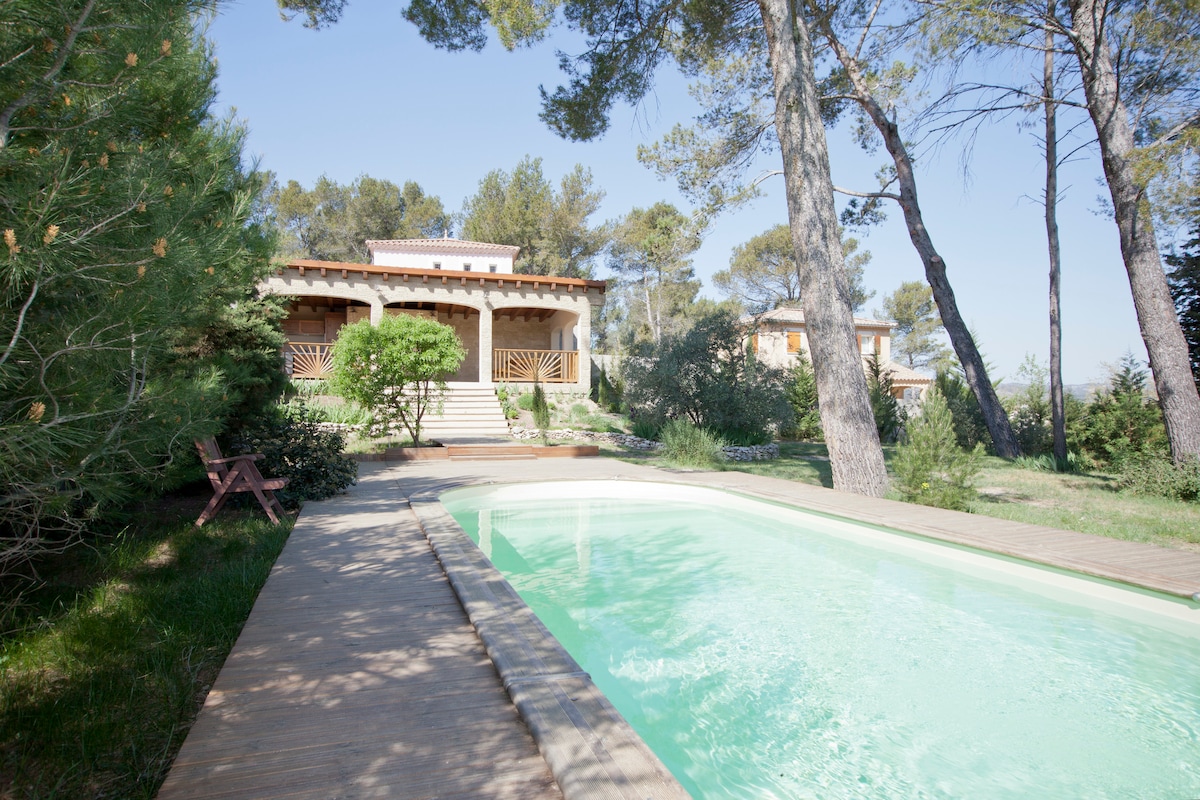 A serene garden setting showcases a refreshing swimming pool bordered by a wooden deck. An inviting house with a covered porch is framed by tall pine trees, with landscaped greenery surrounding the area. Clear blue skies enhance the peaceful atmosphere.